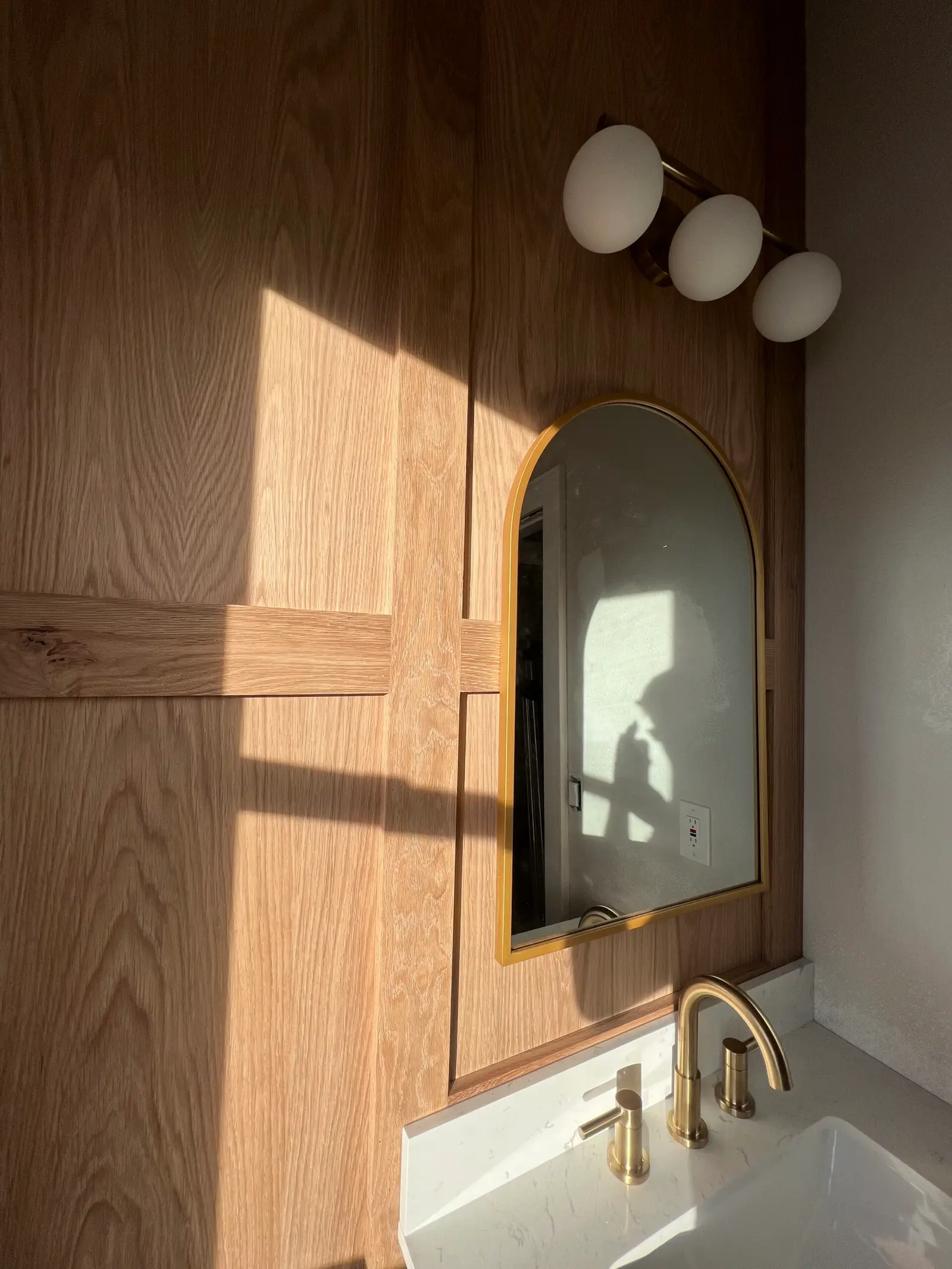 Light wood wall paneling around an arched mirror in a bathroom